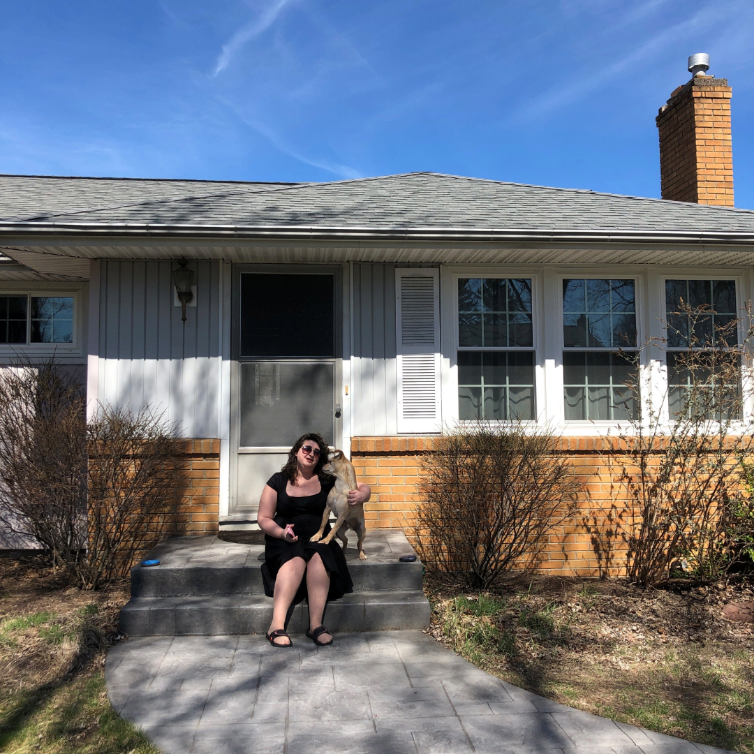 A person sitting on her front steps with a dog in front of a single story house.