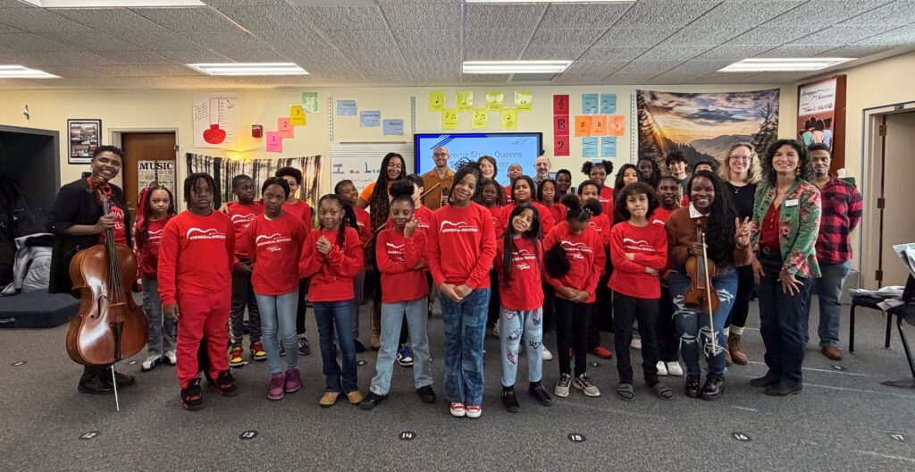 A classroom full of young students in red Strings for Success shirts and a few adults holding musical instruments.