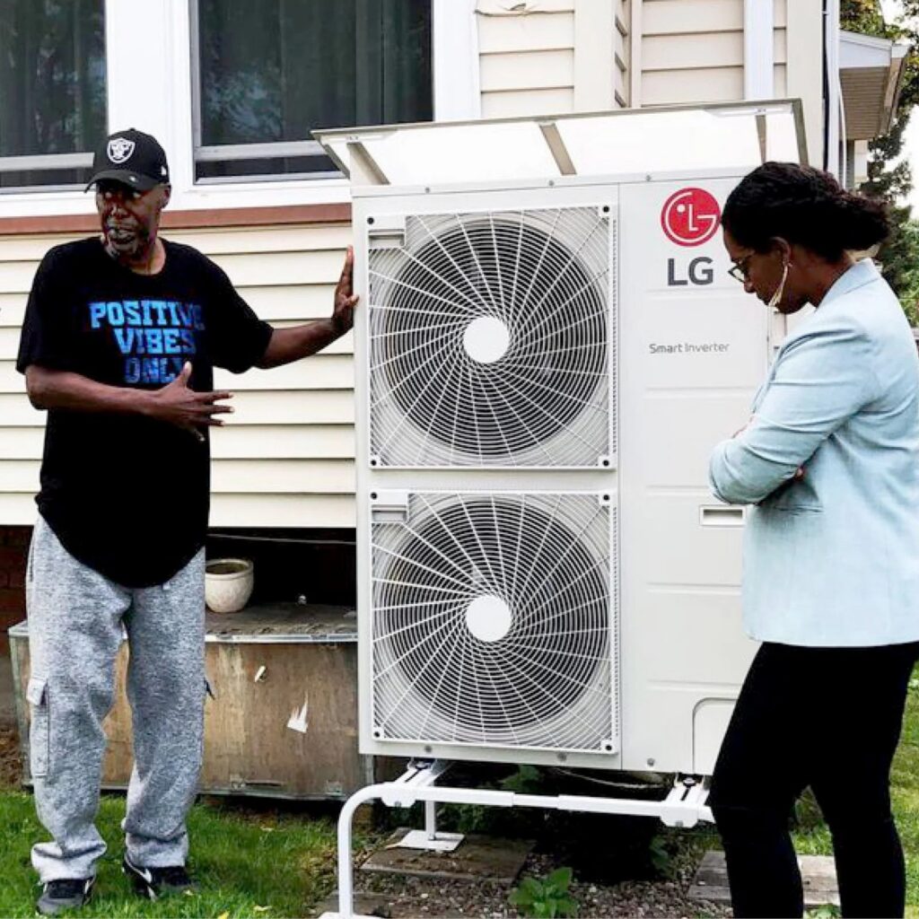 A RENEW client wearing a "positive vibes only" tshirt stands next to Senator Samra Brouk and an outdoor HVAC system.