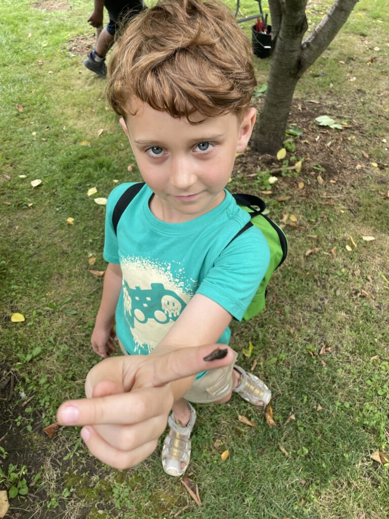 A young boy holds out his finger with a bug on it wearing a teal shirt and a green backpack.
