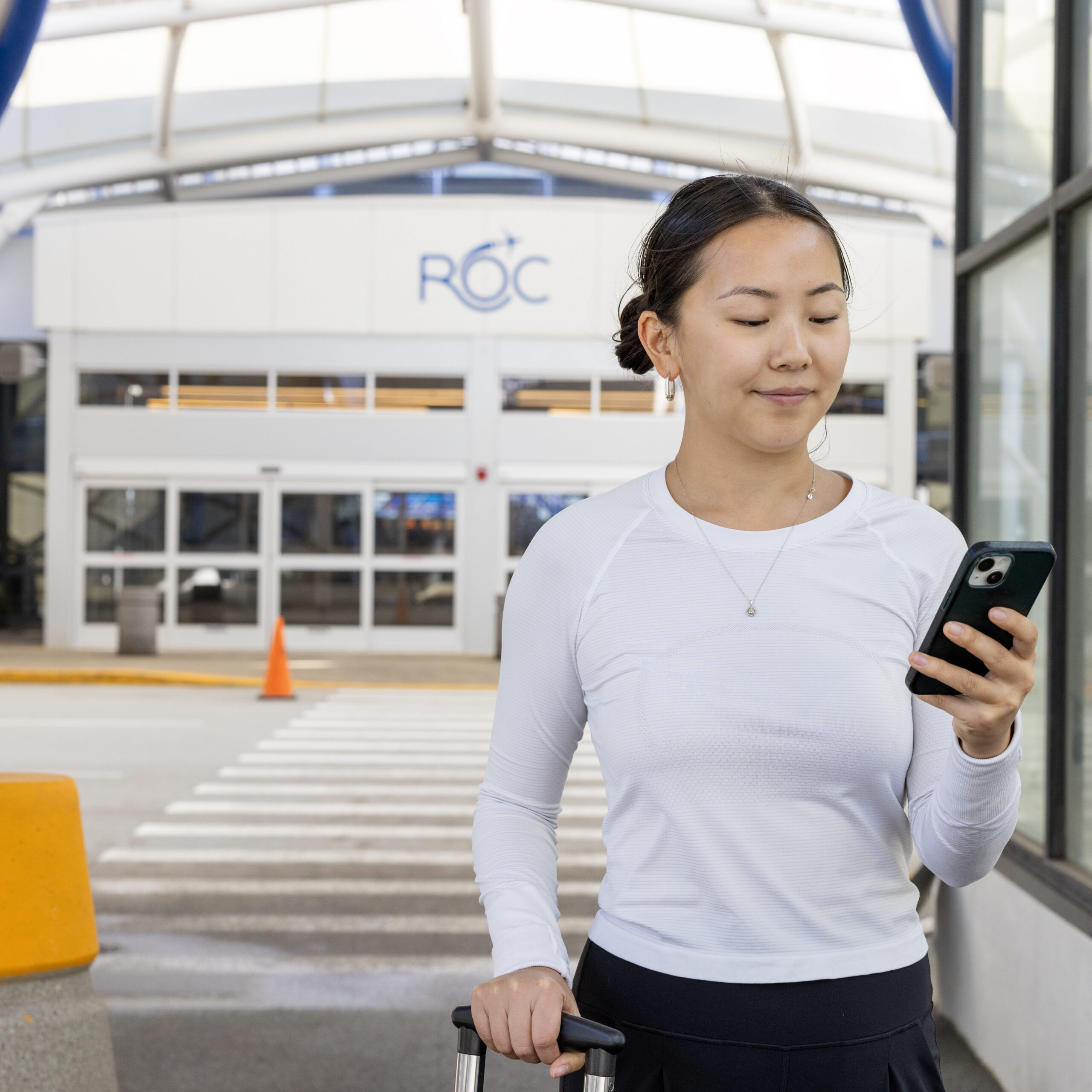 A woman in a white long-sleeved shirt looks at her phone standing in front of an entrance to the Rochester Airport.