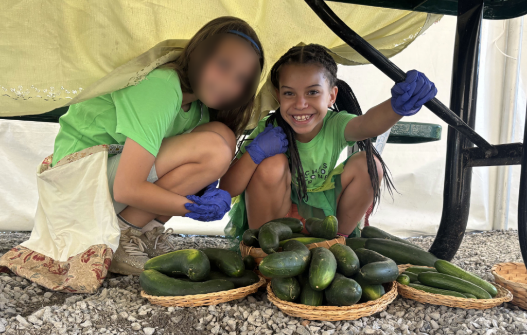 Two young girls crouch next to a large pile of cucmbers.