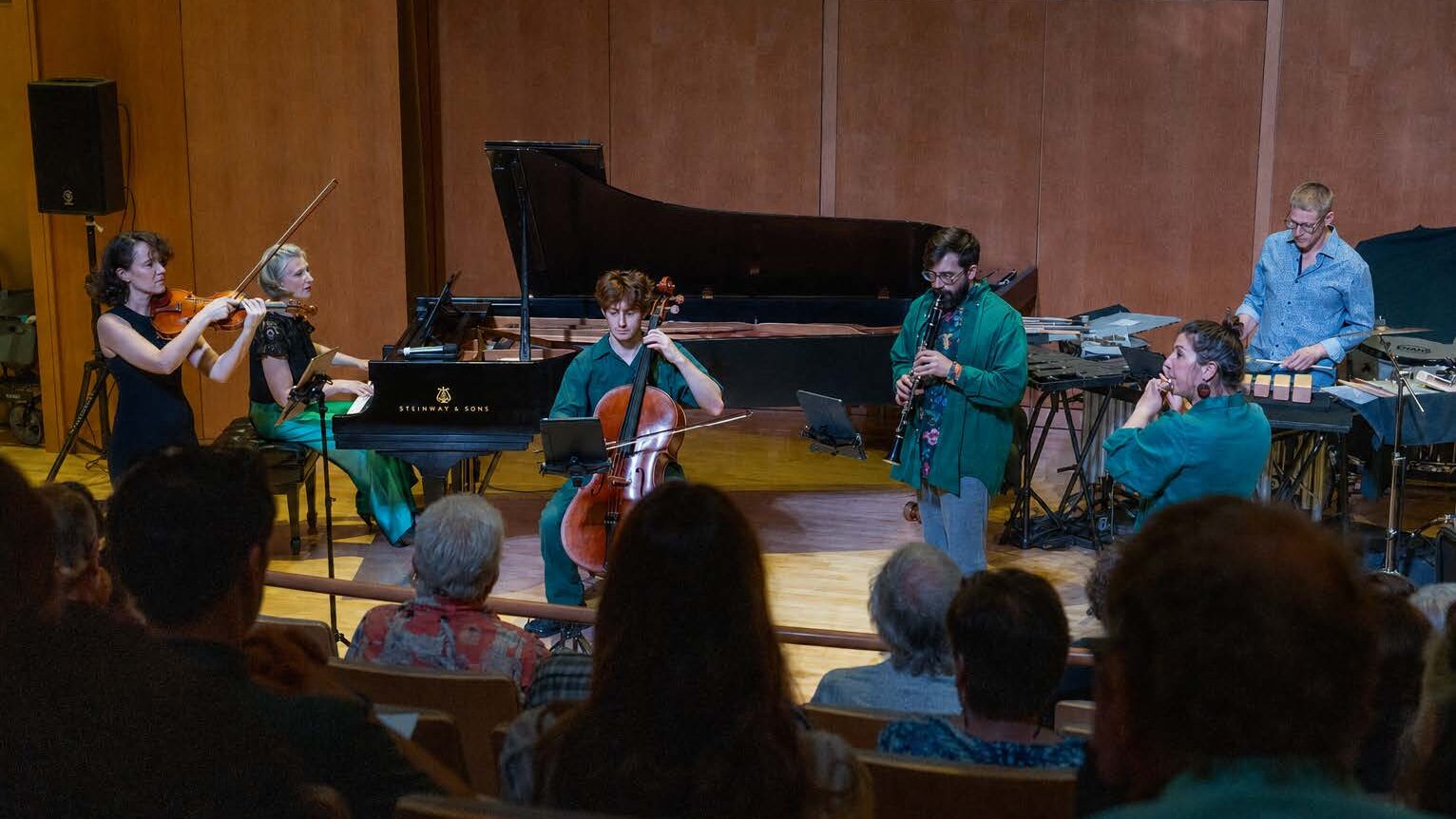 Six musicians on stage playing various instruments with a crowd from behind in the foreground.