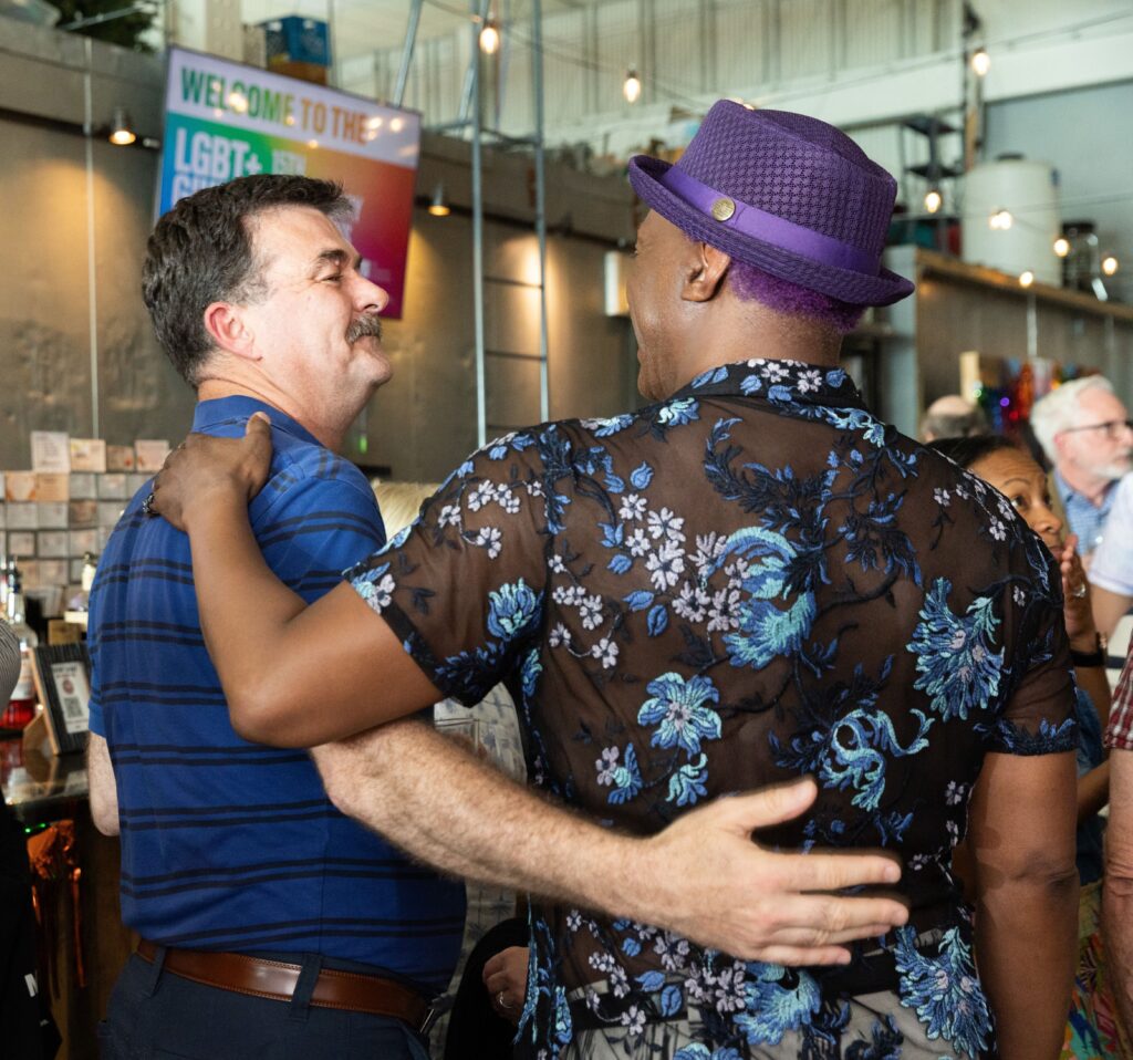 A person in a royal blue and black striped polo with his arm around another person in a black and bright blue floral shirt and a purple hat.