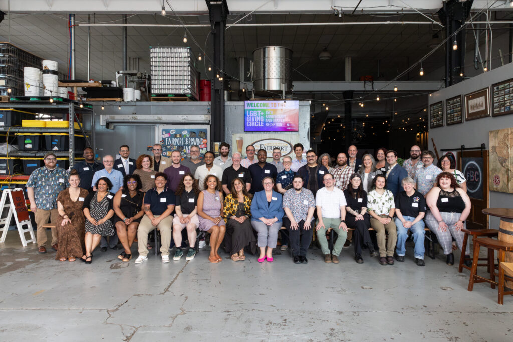 About 40 people sit or stand smiling at the camera in front of a sign that says welcome to the LGBT+ Giving Circle 15th Anniversary and grants reception.
