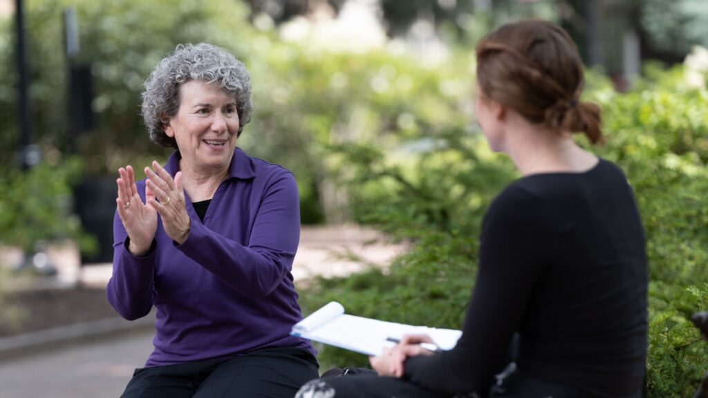 A person with curly hair in a purple top gestures and talks to someone holding a pen and paper sitting outside the Memorial Art Gallery.