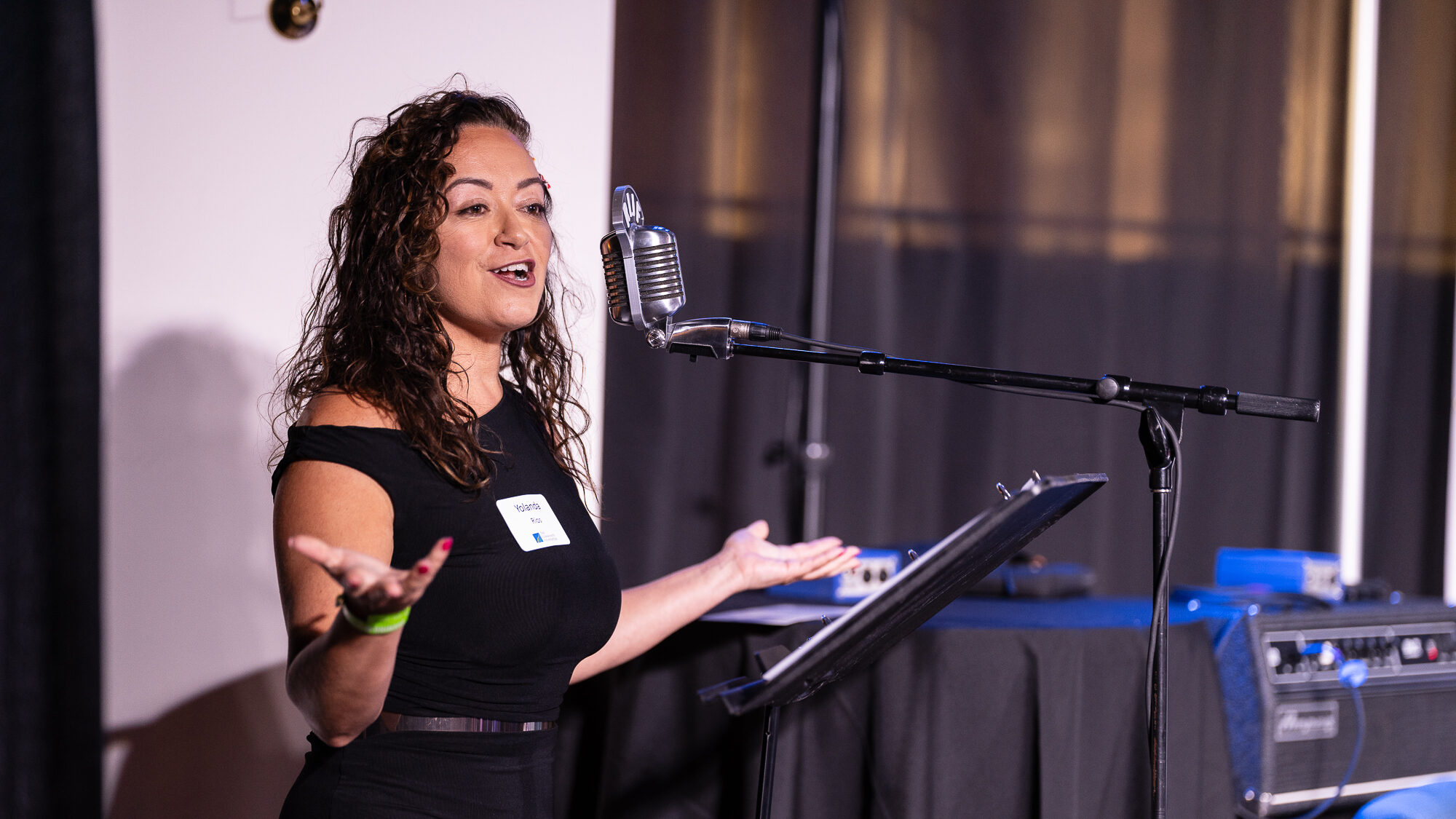 Yolanda Rios in black evening wear gesturing with hands outstretched at a podium.