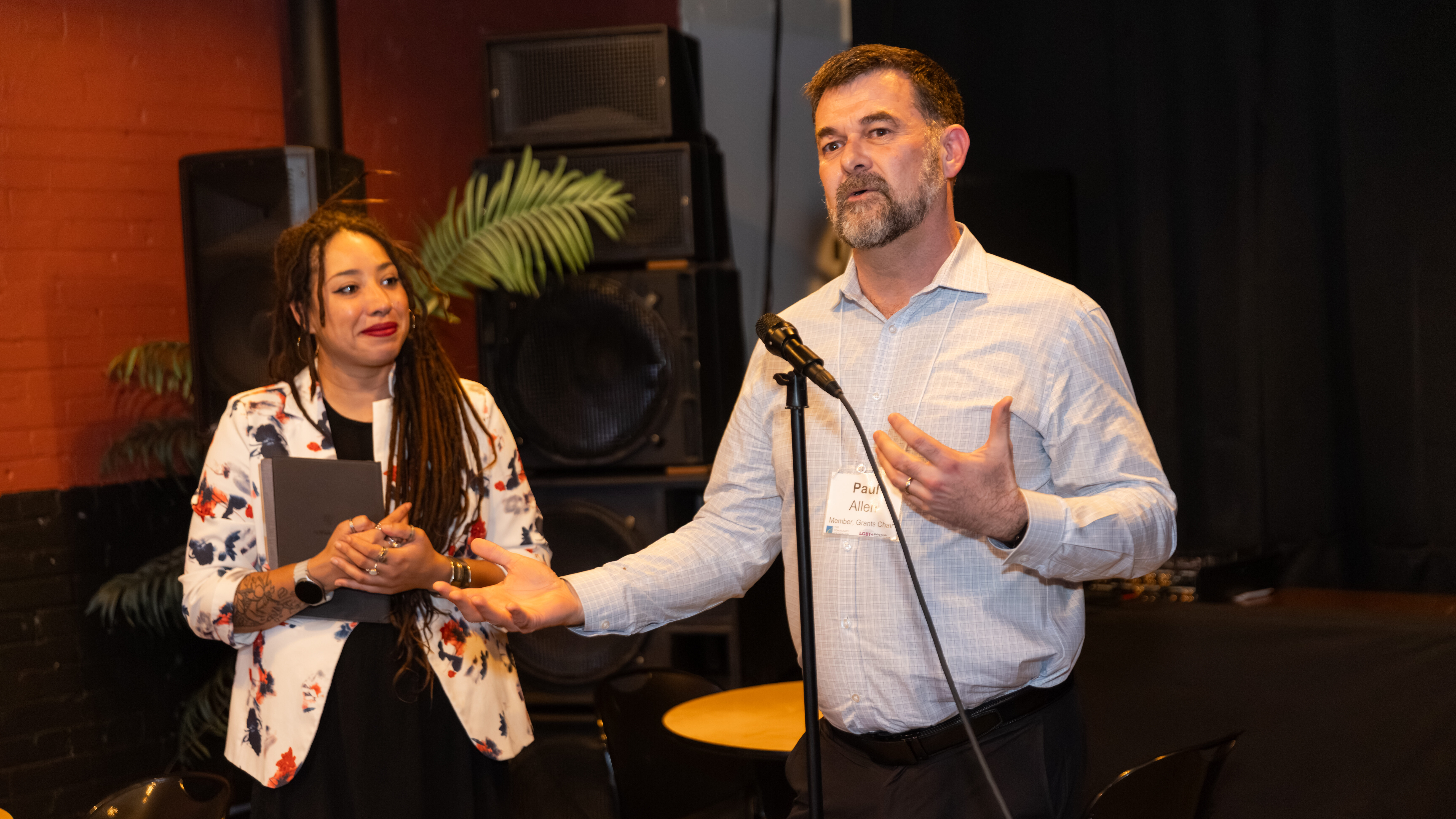 Dawn Perry, in a white jacket with blue and orange flowers, looks thoughtfully at a man speaking at a microphone.