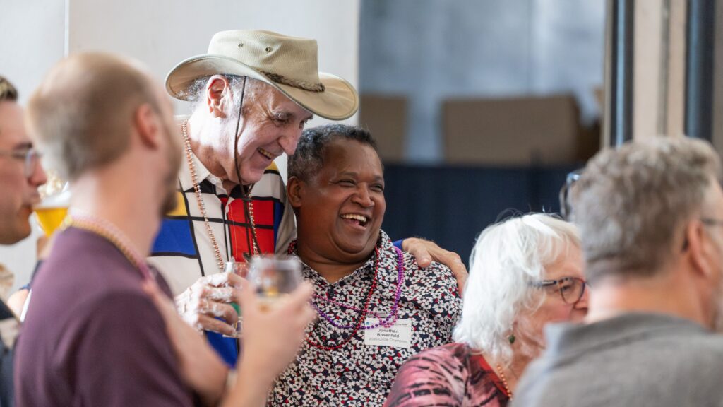 A man in a beige hat and colorful top hugs another man in a floral shirt from the side.