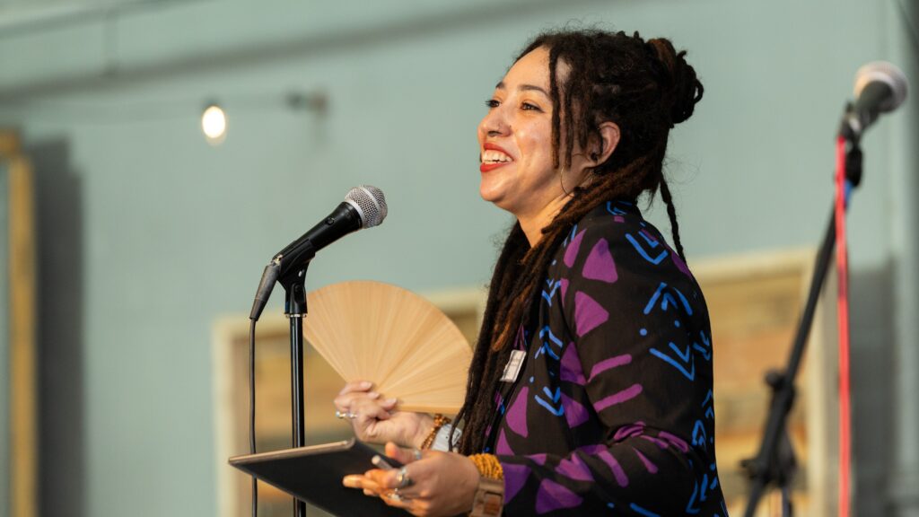 Someone in a black jacket with purple and blue designs holds a fan and smiles in front of a microphone.