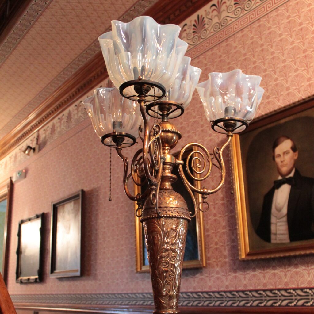 An ornate sconce on the railing in a hallway with wood paneling, pink wallpaper and portraits.