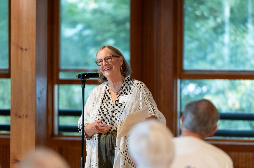 Nancy Rees in a white and black top and green pants smiles in front of a microphone at Camp Cory.